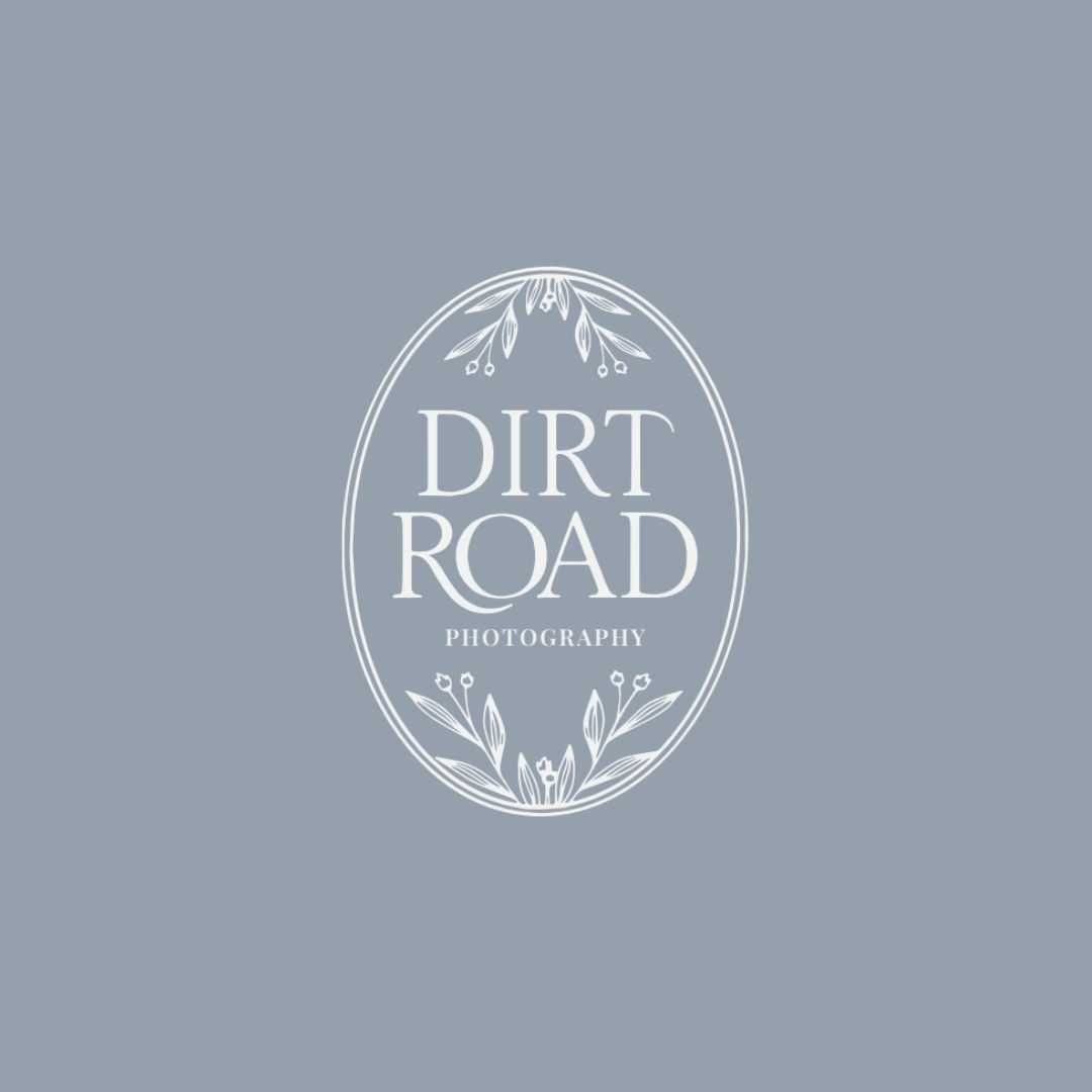 Minimal logo design for Dirt Road Photography with white serif typography inside an oval floral frame on a muted blue background, created for a storytelling focused family and senior photography brand by Draftss